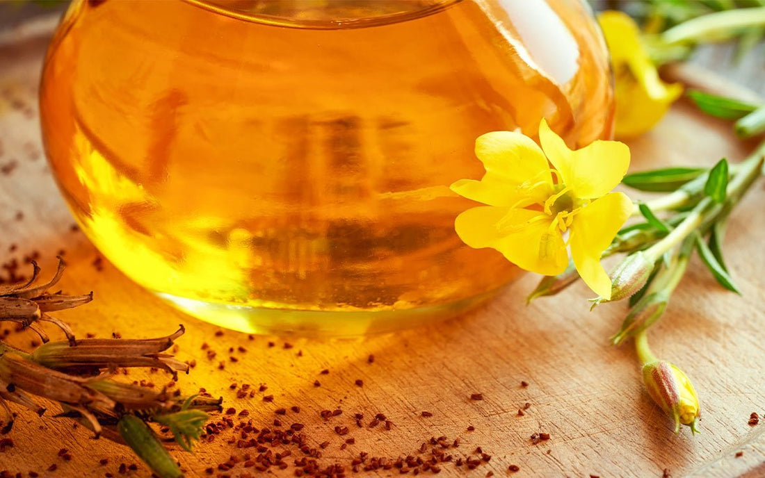 Ein Glasgefäß mit Öl gefüllt, Blüten der Nachtkerze und Pflanzensamen auf einem Holzbrett verstreut